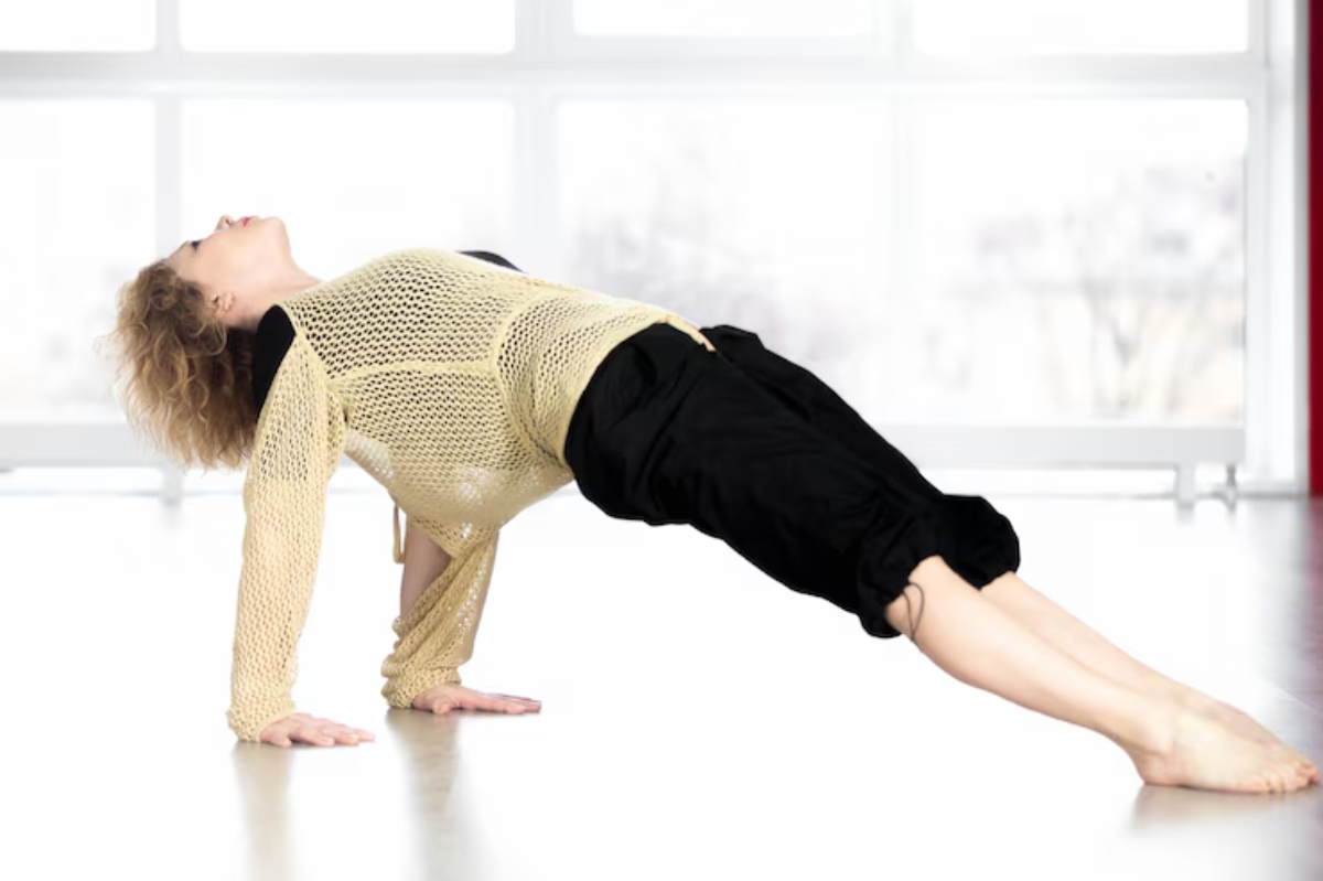 A person in a sheer, beige sweater and black pants performs a yoga pose on a wooden studio floor, with large windows in the background.