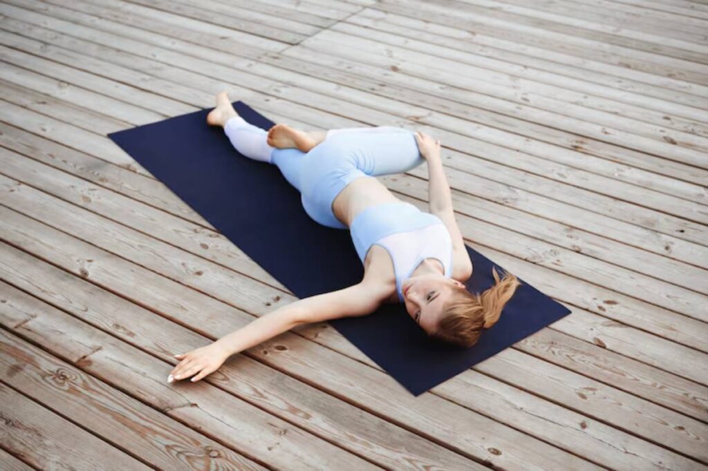A woman in light blue activewear performs a yoga stretch on a black mat, set against a wooden deck surface.