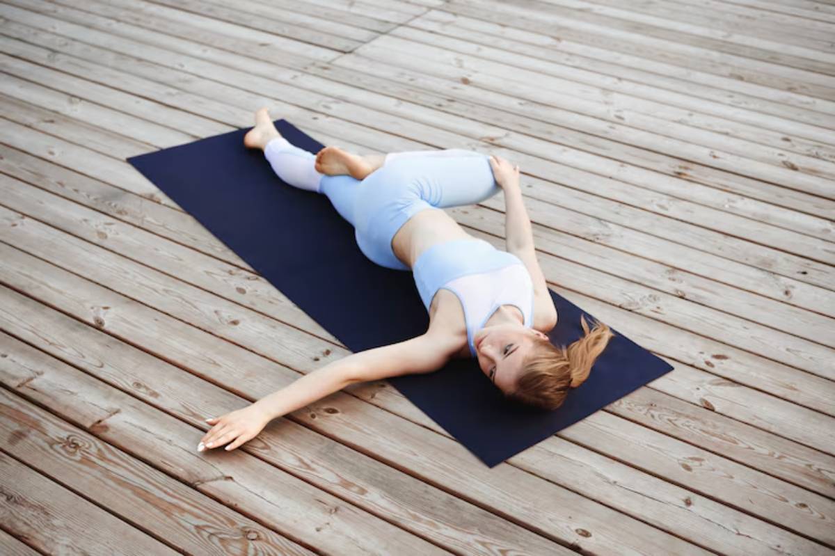 A woman in light blue activewear performs a yoga stretch on a black mat, set against a wooden deck surface.