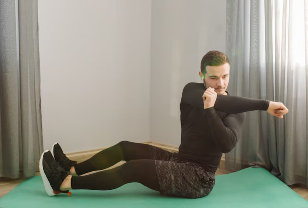 A person in workout attire performs a seated exercise on a green mat, with natural light illuminating the room.