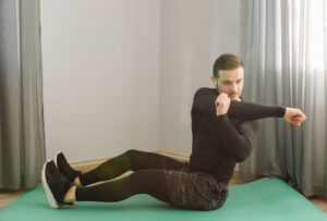 A person in workout attire performs a seated exercise on a green mat, with natural light illuminating the room.