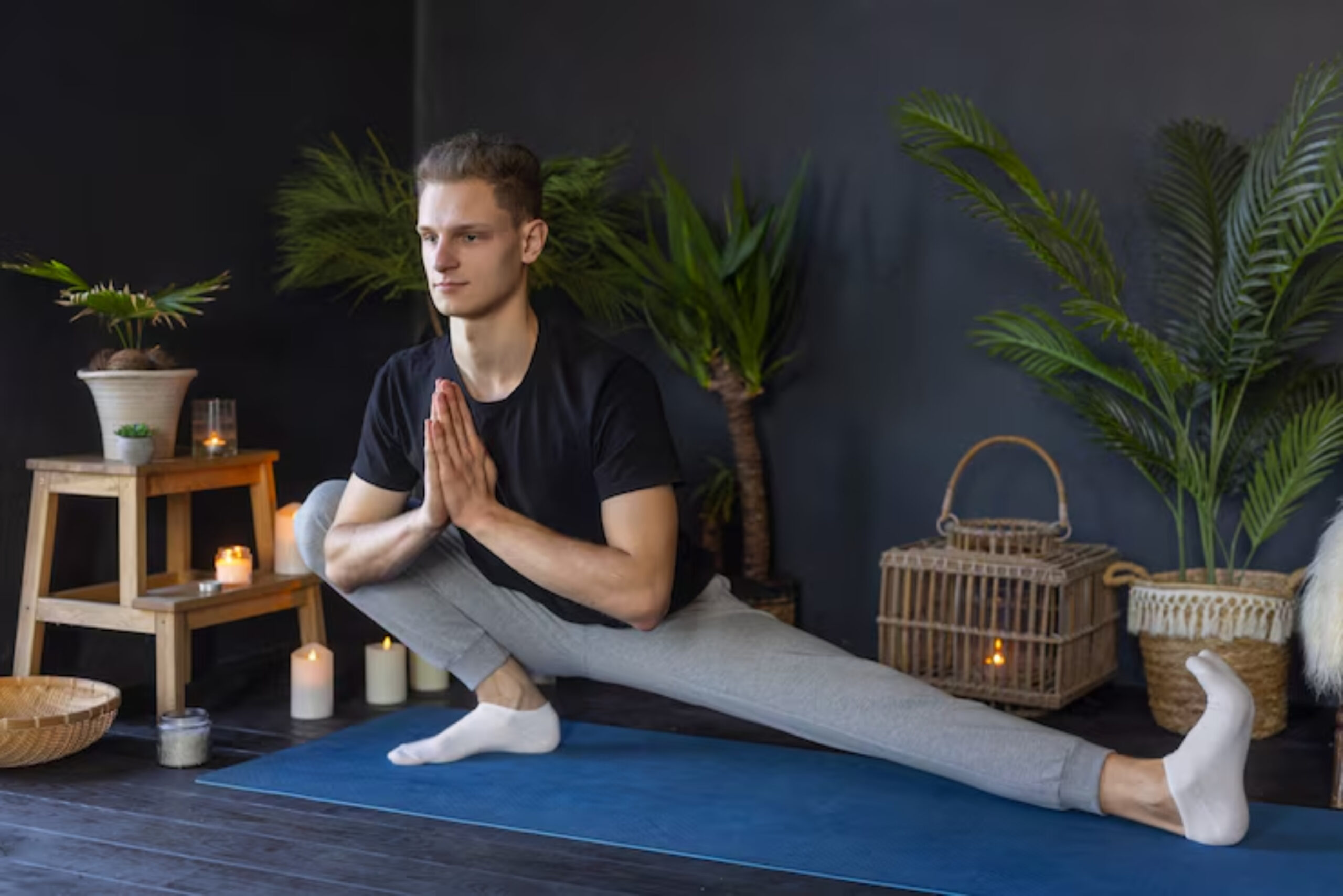 A person in a yoga pose on a blue mat surrounded by plants, candles, and decorative baskets in a serene indoor setting. 