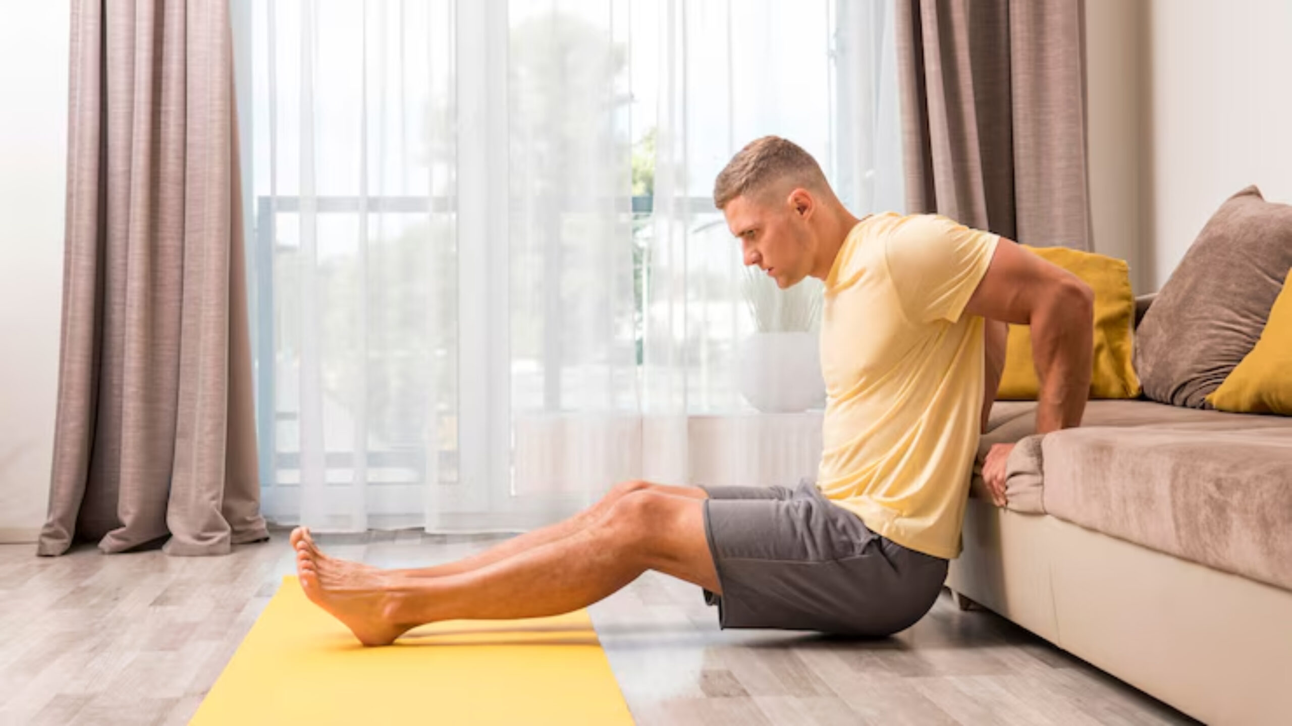 A person in a yellow shirt performs tricep dips on the edge of a couch, using a yoga mat in a well-lit living room setting. 