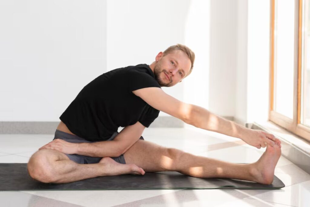 A person practicing yoga, seated on a mat, stretches forward to reach their toes in a sunlit room with large windows.