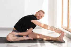 A person practicing yoga, seated on a mat, stretches forward to reach their toes in a sunlit room with large windows.