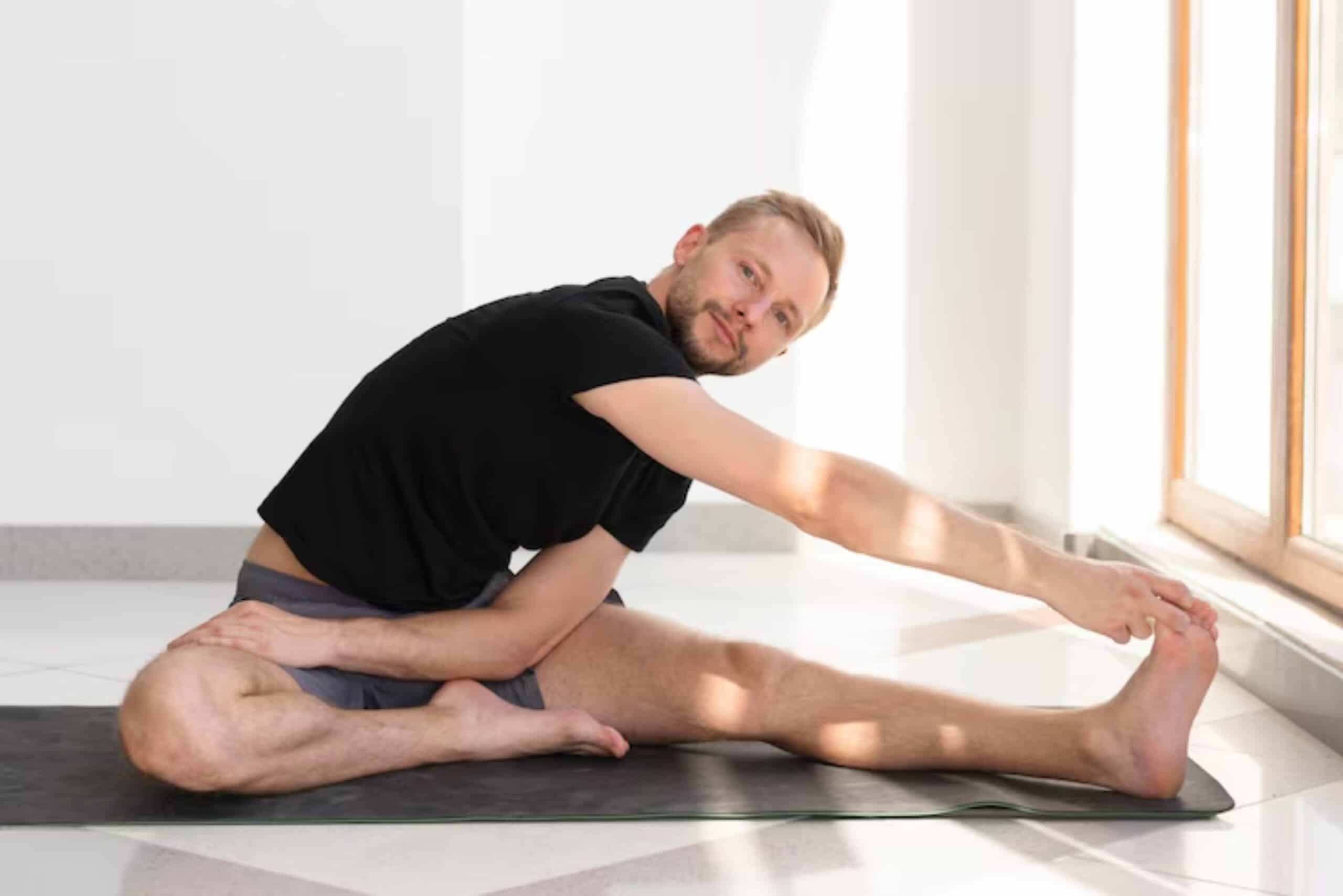 A person practicing yoga, seated on a mat, stretches forward to reach their toes in a sunlit room with large windows.