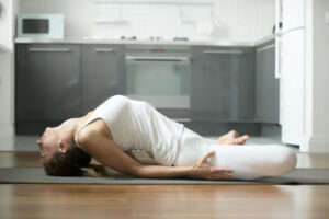 A woman in a white outfit performs a yoga pose on a mat in a modern kitchen, showcasing flexibility and tranquility.