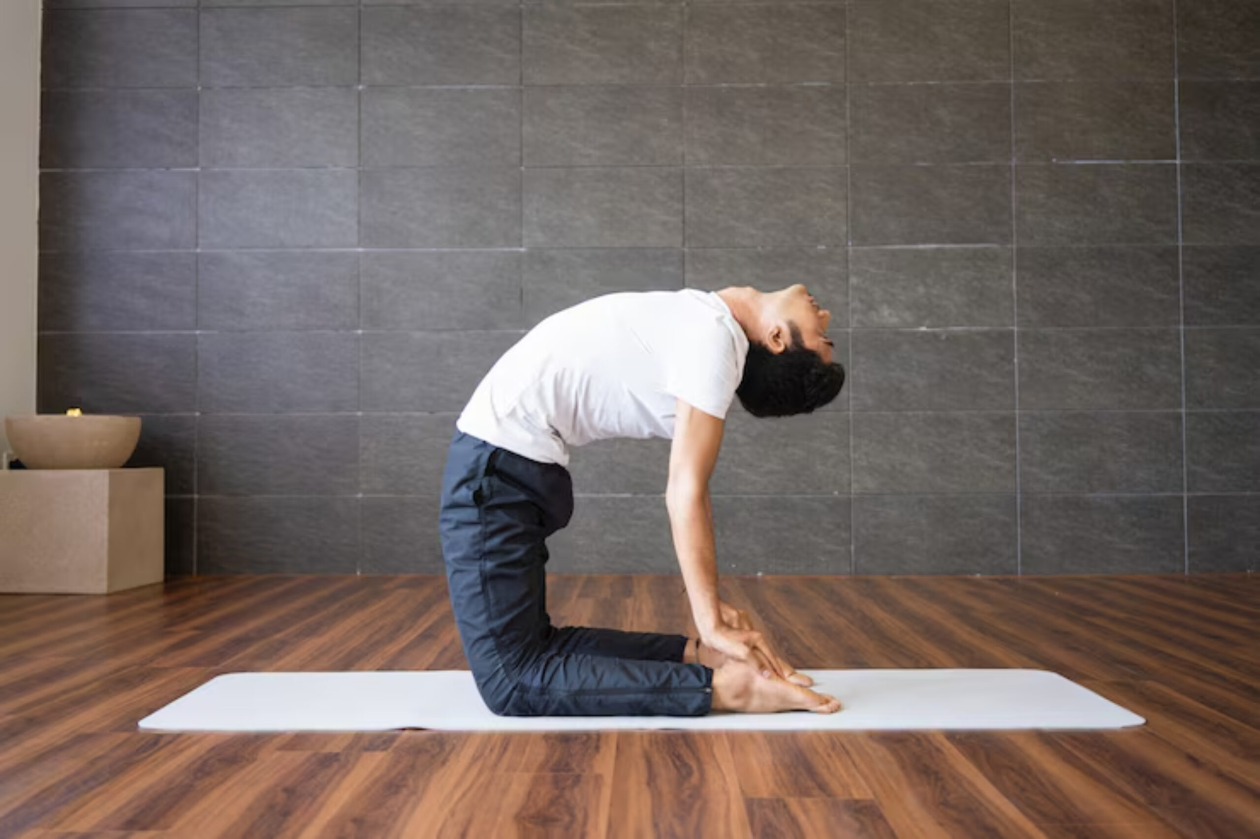 A person performs a yoga camel pose on a mat, stretching backward with hands on their heels, in a serene indoor space.