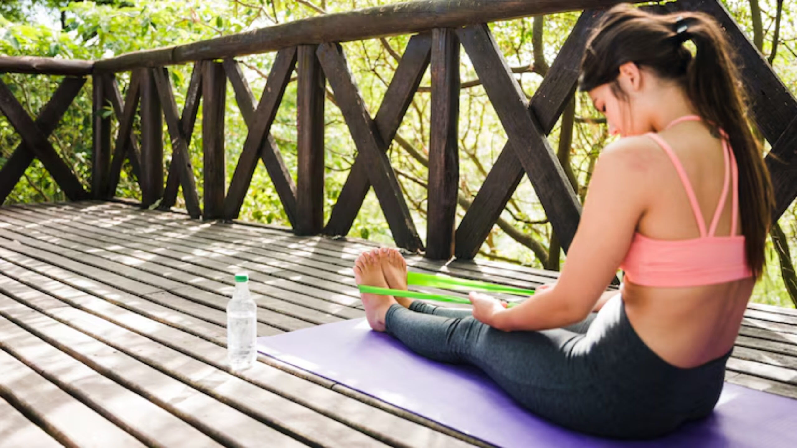  A person sits on a yoga mat on a wooden deck surrounded by greenery, using a resistance band to stretch their feet. A water bottle rests nearby.