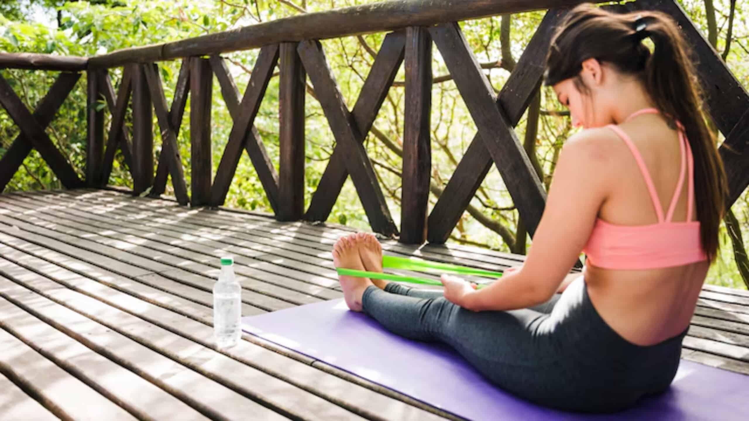 A person sits on a yoga mat on a wooden deck surrounded by greenery, using a resistance band to stretch their feet. A water bottle rests nearby.