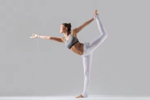 A woman in a gray sports bra and white leggings performs a yoga pose, balancing gracefully on one leg with arms outstretched.