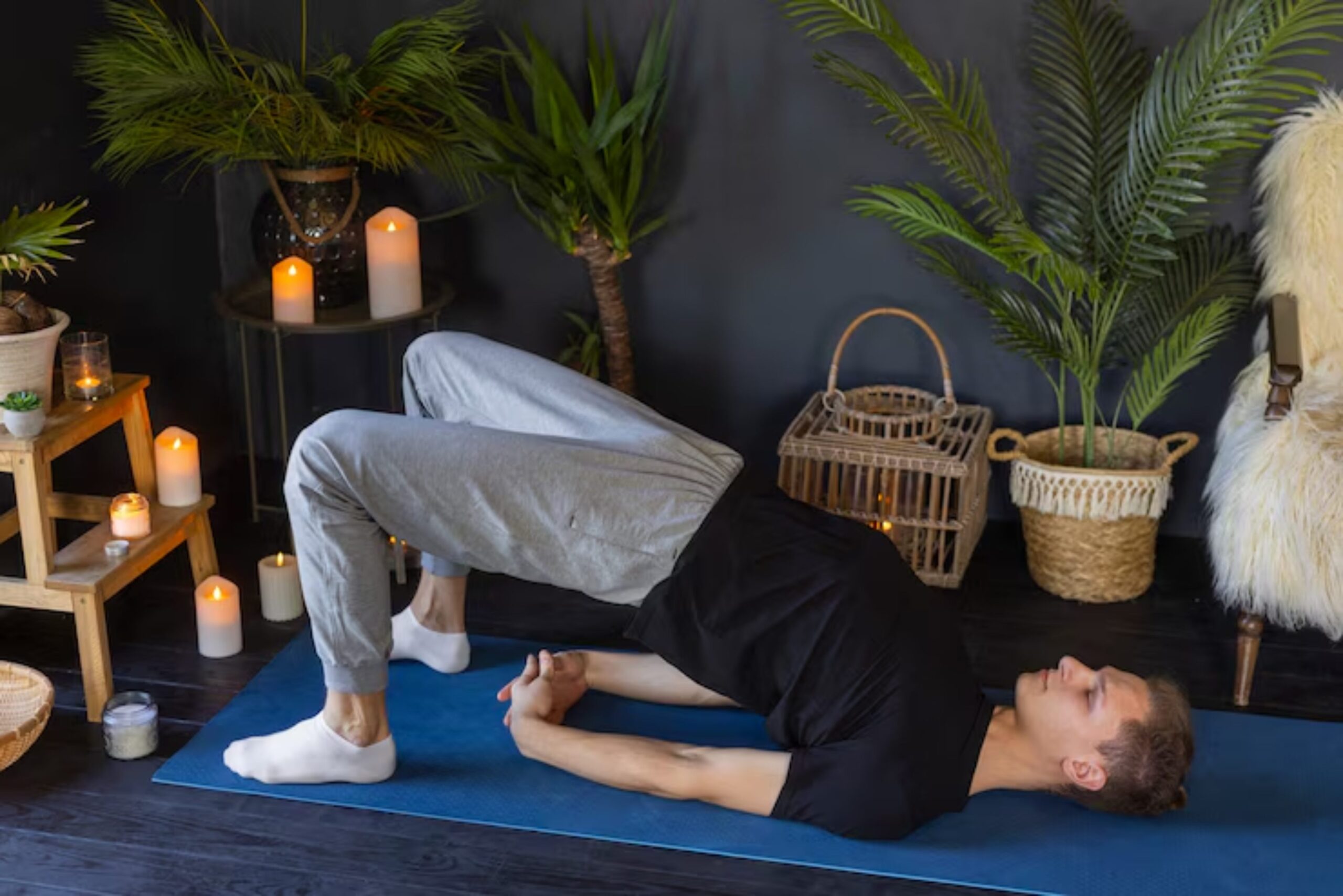 A person practices yoga on a blue mat surrounded by candles and tropical plants in a serene indoor setting. 