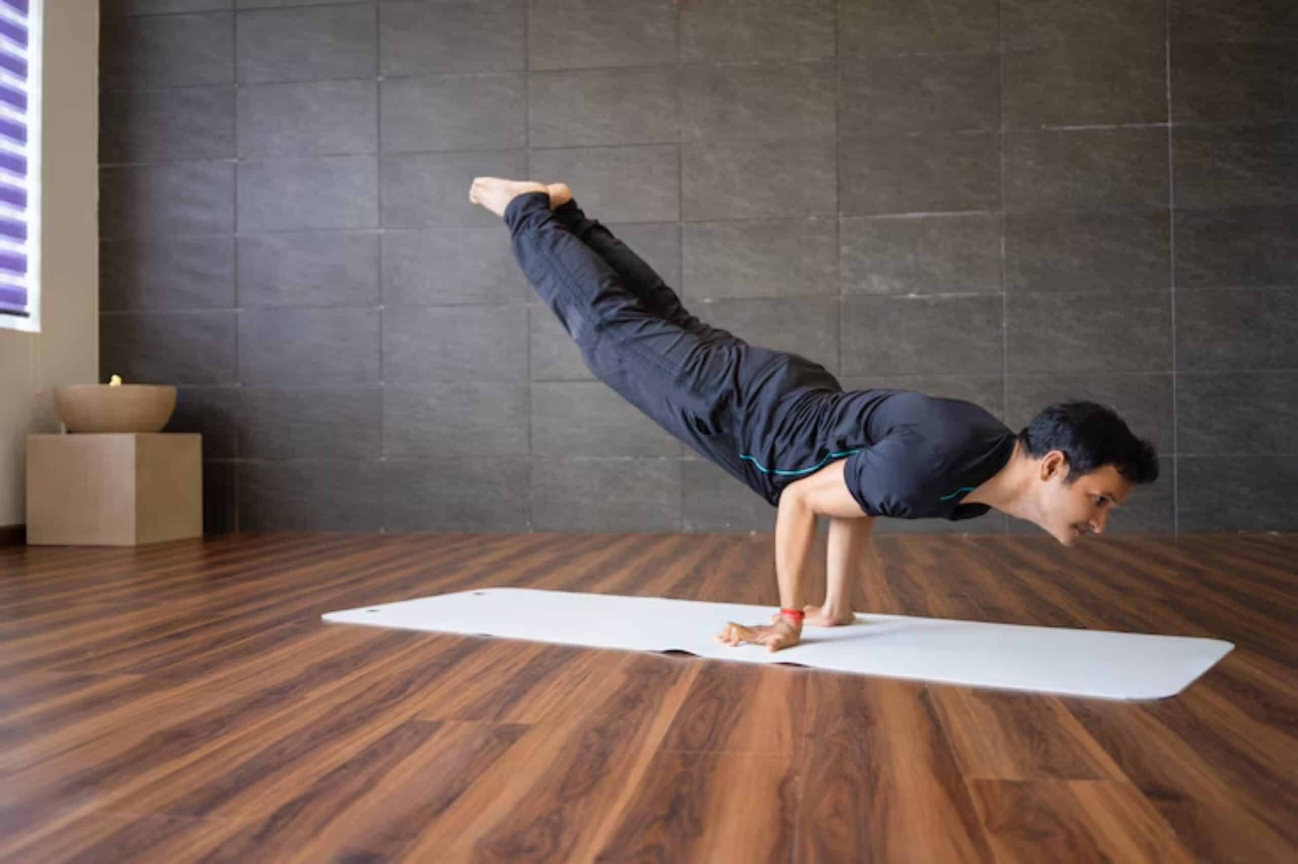 A person performs a challenging yoga pose on a white mat, balancing on hands with legs extended upward in a spacious, minimalist studio.