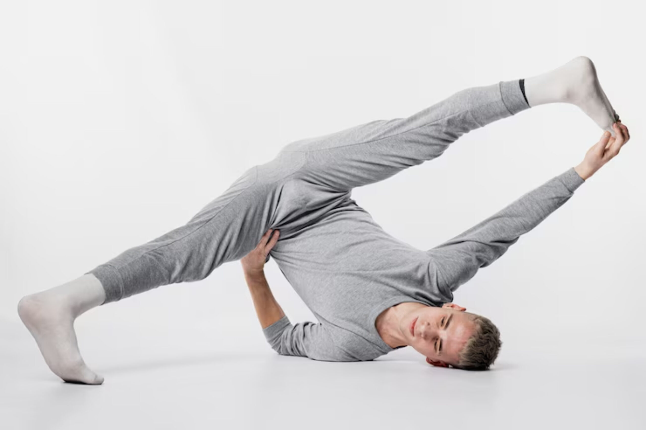 A person in gray loungewear performs an acrobatic stretch, balancing on one arm while extending a leg overhead. 