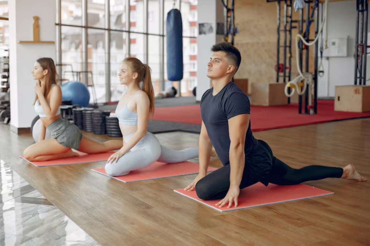 Three individuals practice yoga on mats in a bright gym, focusing on stretching and flexibility exercises.