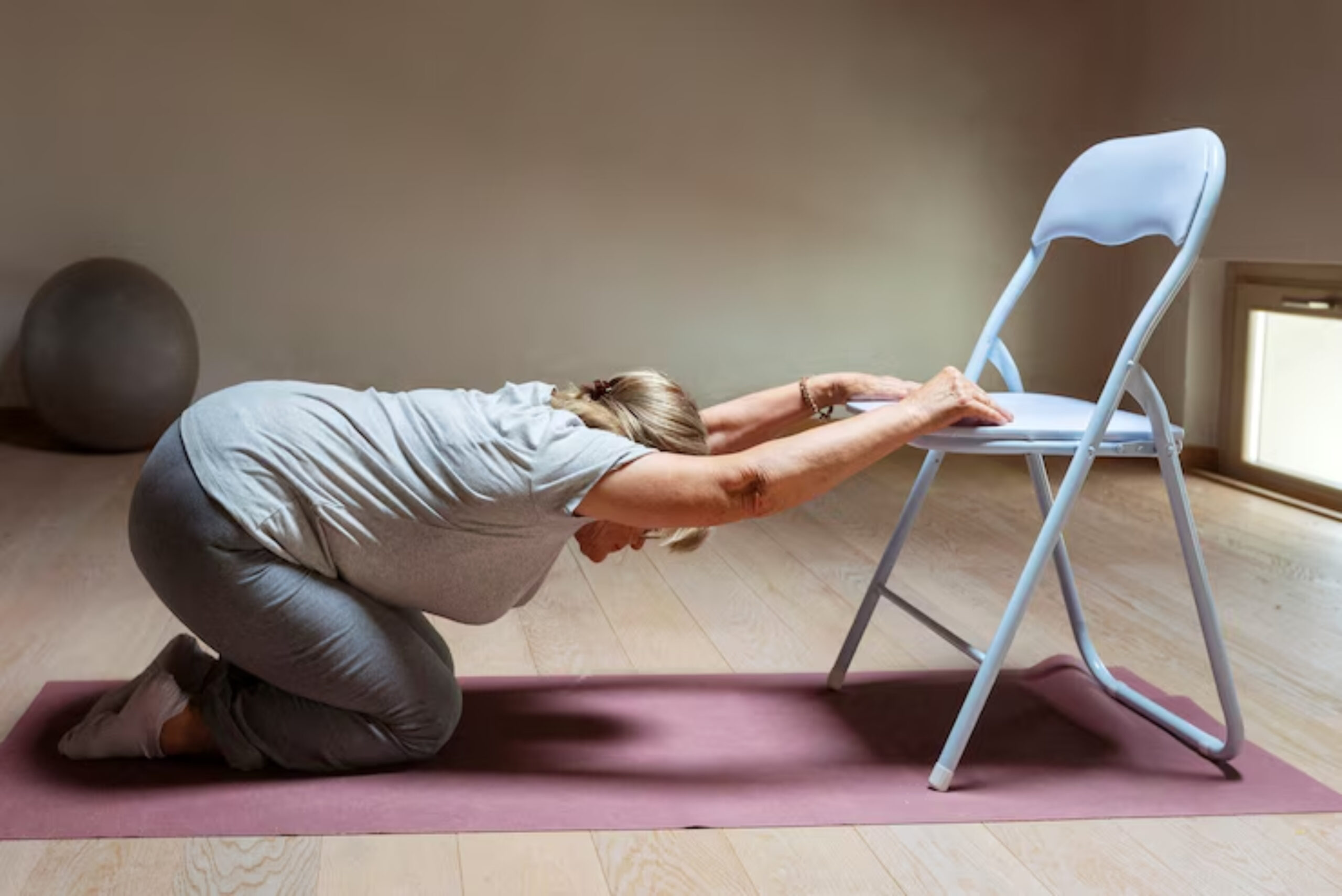 An elderly woman in a gray outfit performs a stretching exercise using a chair for support on a yoga mat. 