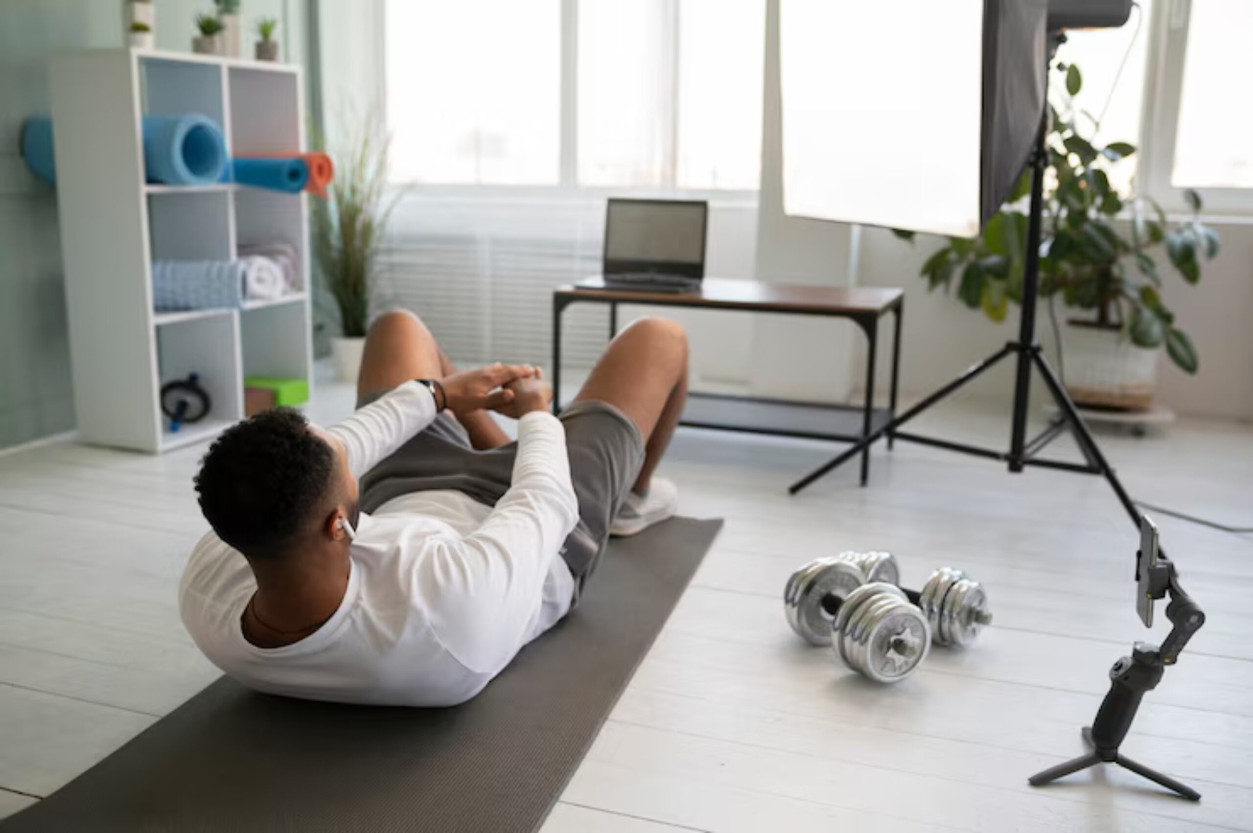 A young man exercises on a mat at home, with dumbbells nearby and a laptop set up for a workout session. 