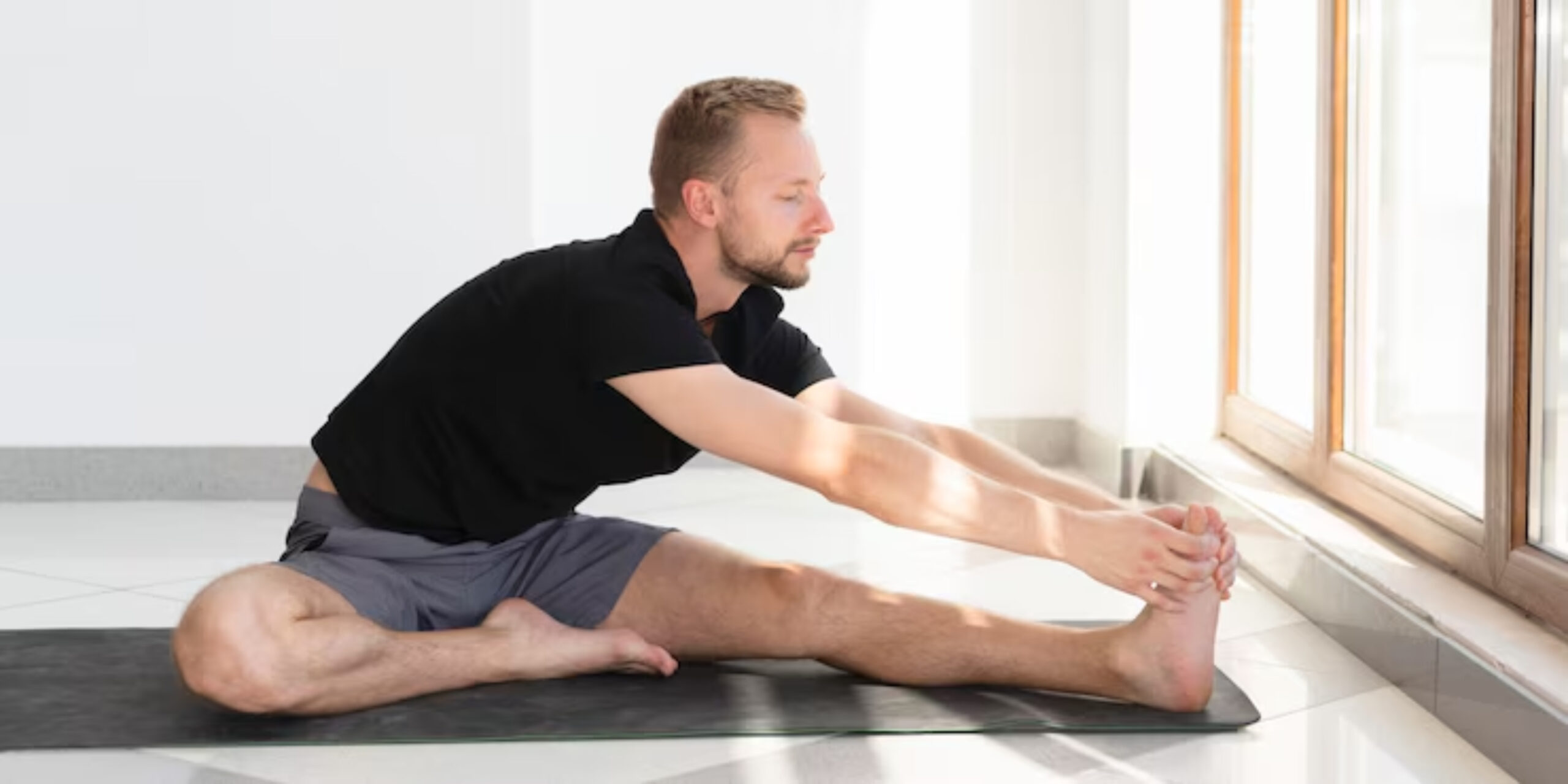 A person is stretching on a yoga mat, reaching for their foot, with sunlight filtering through large windows. 