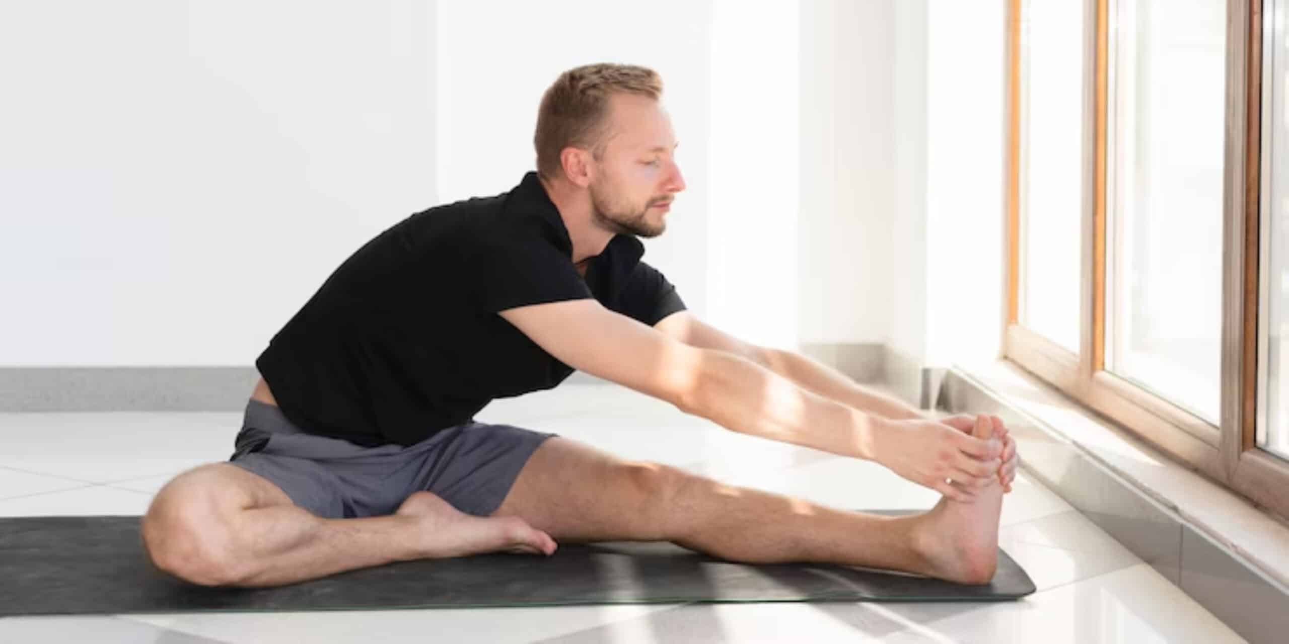 A person is stretching on a yoga mat, reaching for their foot, with sunlight filtering through large windows.