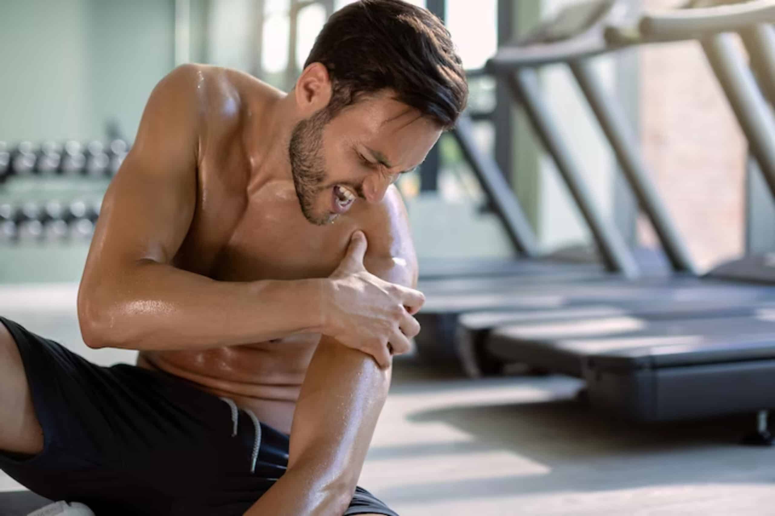 A man in athletic wear sits on the gym floor, massaging his shoulder, with sweat glistening on his body amid workout equipment.
