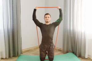 A person in a fitted black shirt and shorts performs an overhead exercise with a red resistance band on a green mat indoors.