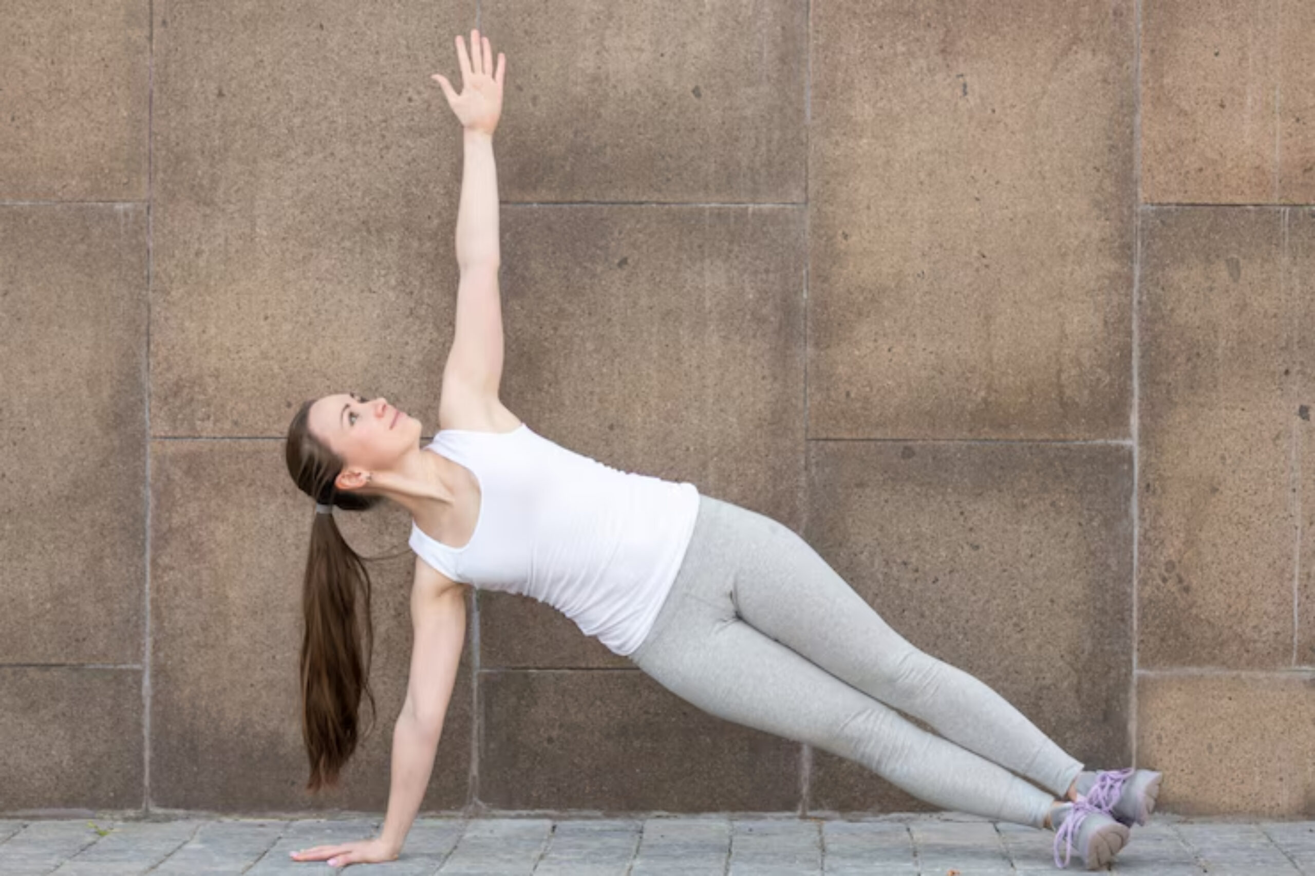 A woman performs a side plank exercise against a stone wall, one arm raised, wearing a white tank top and gray leggings.