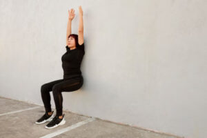 A person in black athletic wear is performing a wall sit against a light-colored wall, arms raised overhead, on a concrete surface.