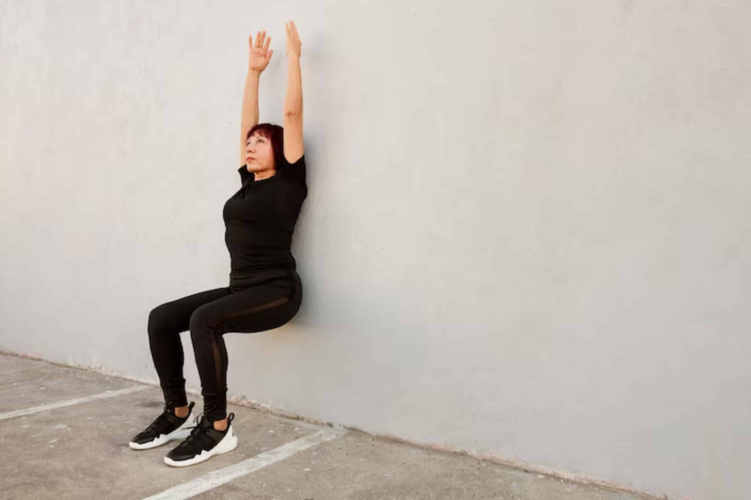 A person in black athletic wear is performing a wall sit against a light-colored wall, arms raised overhead, on a concrete surface.