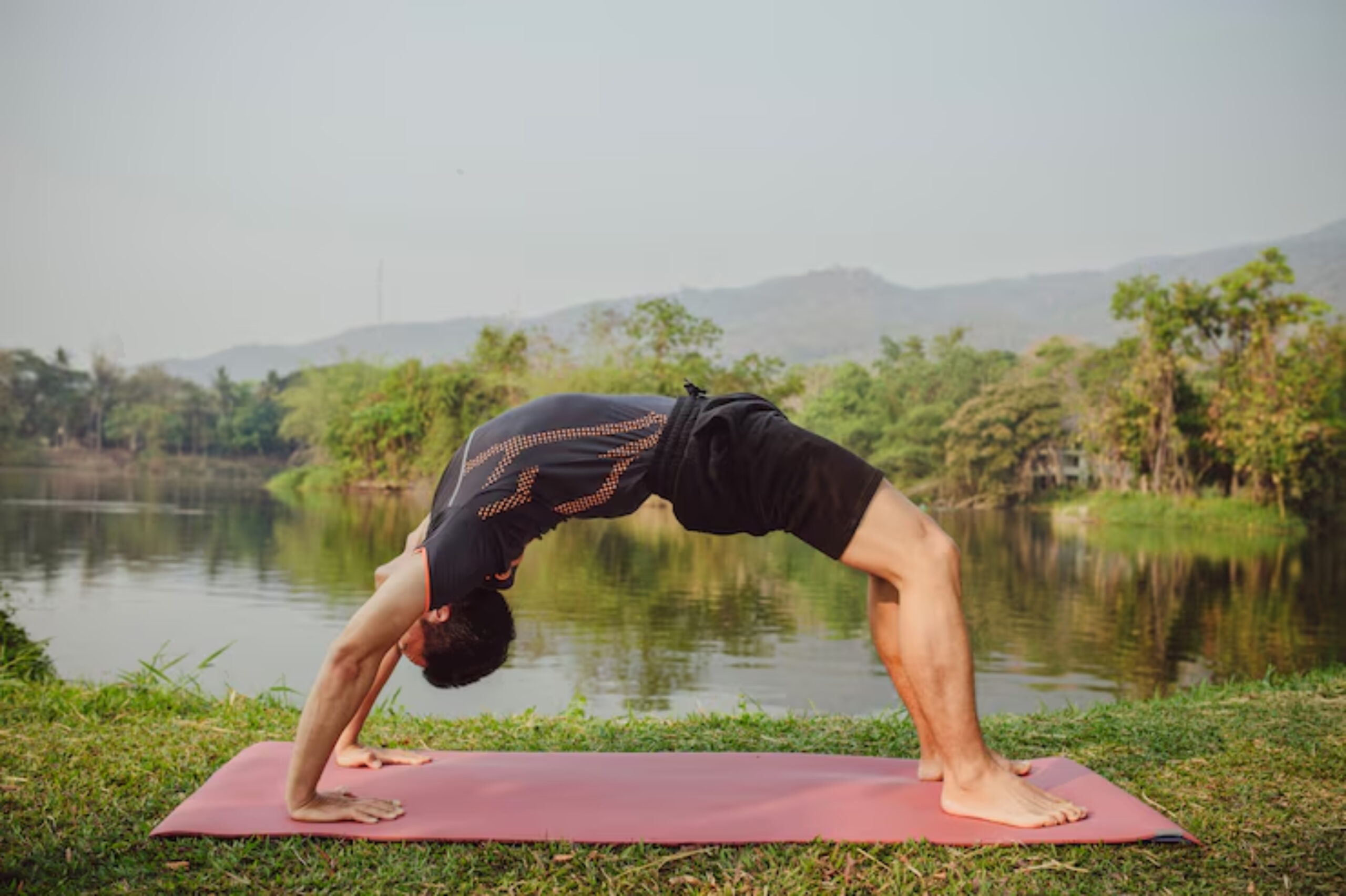 A man performs a backbend yoga pose on a mat by a serene lake, surrounded by lush greenery and mountains in the background. 
