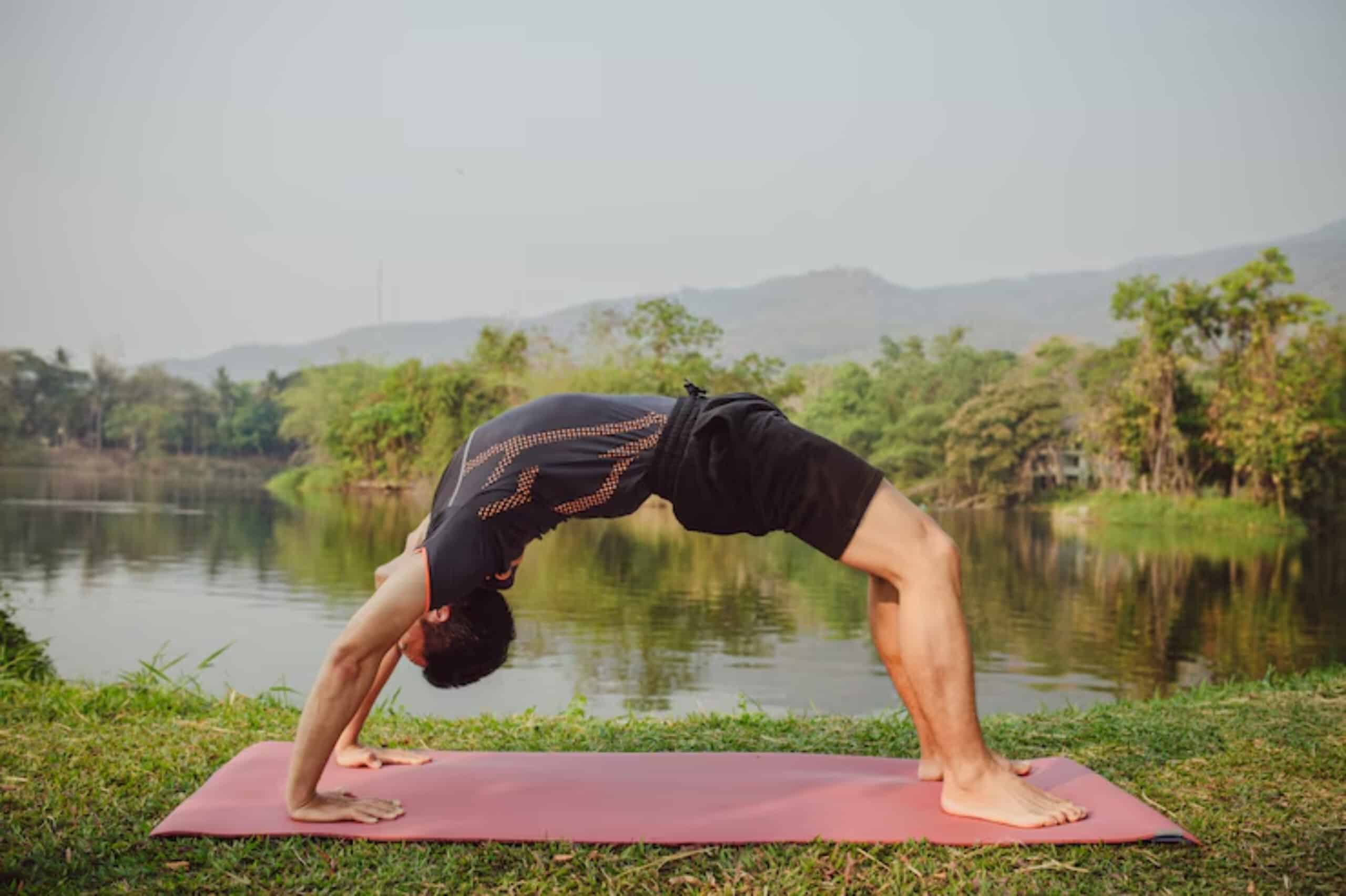 A man performs a backbend yoga pose on a mat by a serene lake, surrounded by lush greenery and mountains in the background.