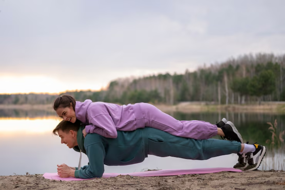 Two individuals in colorful workout attire perform a plank exercise on a yoga mat by a serene lake at sunset.