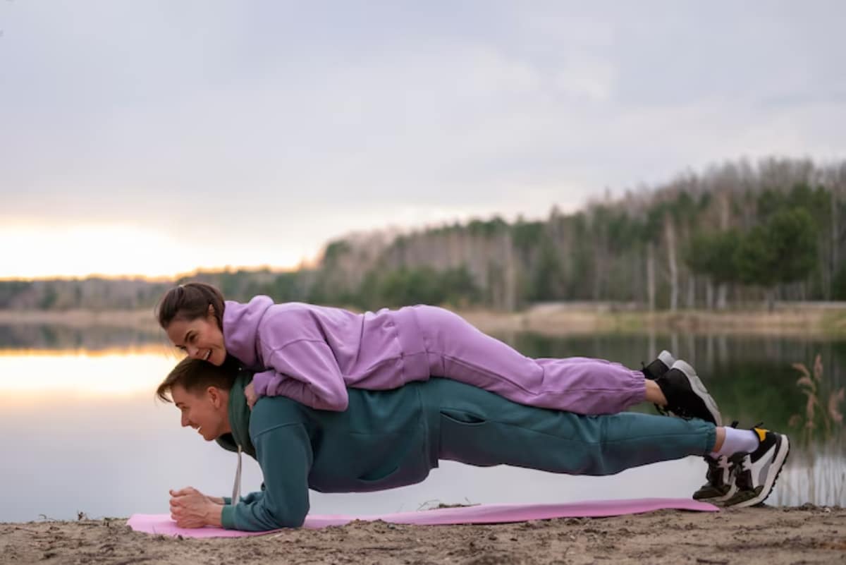 Two individuals in colorful workout attire perform a plank exercise on a yoga mat by a serene lake at sunset.