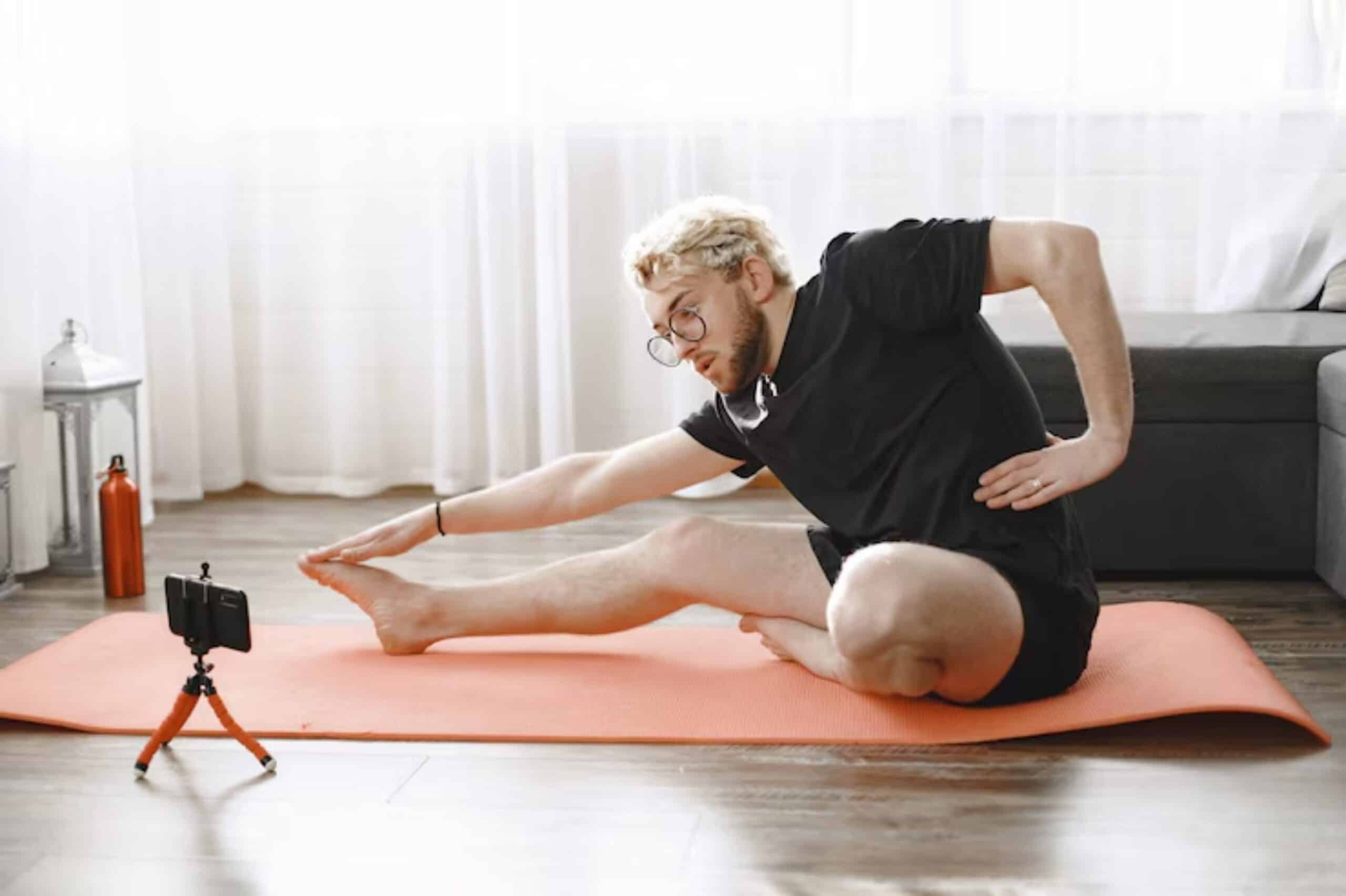 A person in a black shirt stretches on an orange yoga mat at home, while a smartphone on a tripod captures the session.