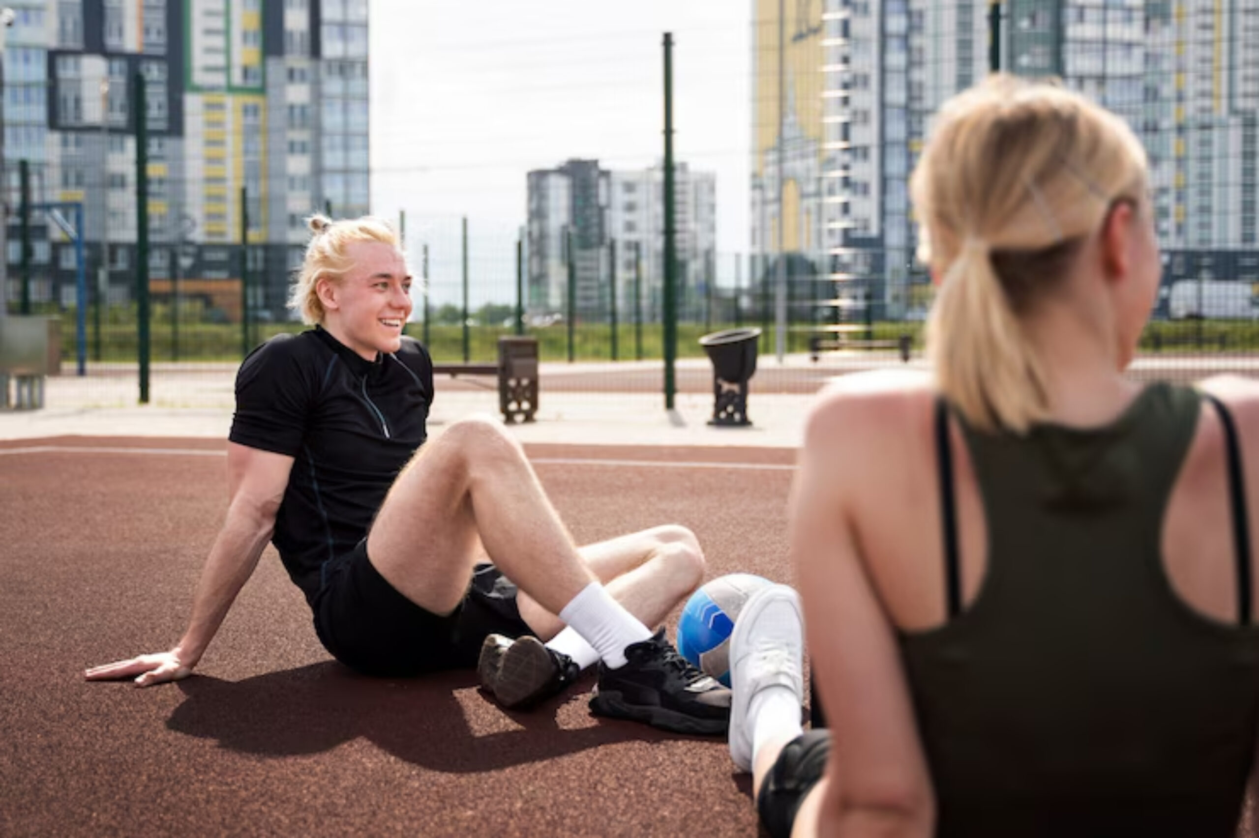 Two individuals seated on a track in an urban outdoor sports facility, with buildings and a fence in the background. 