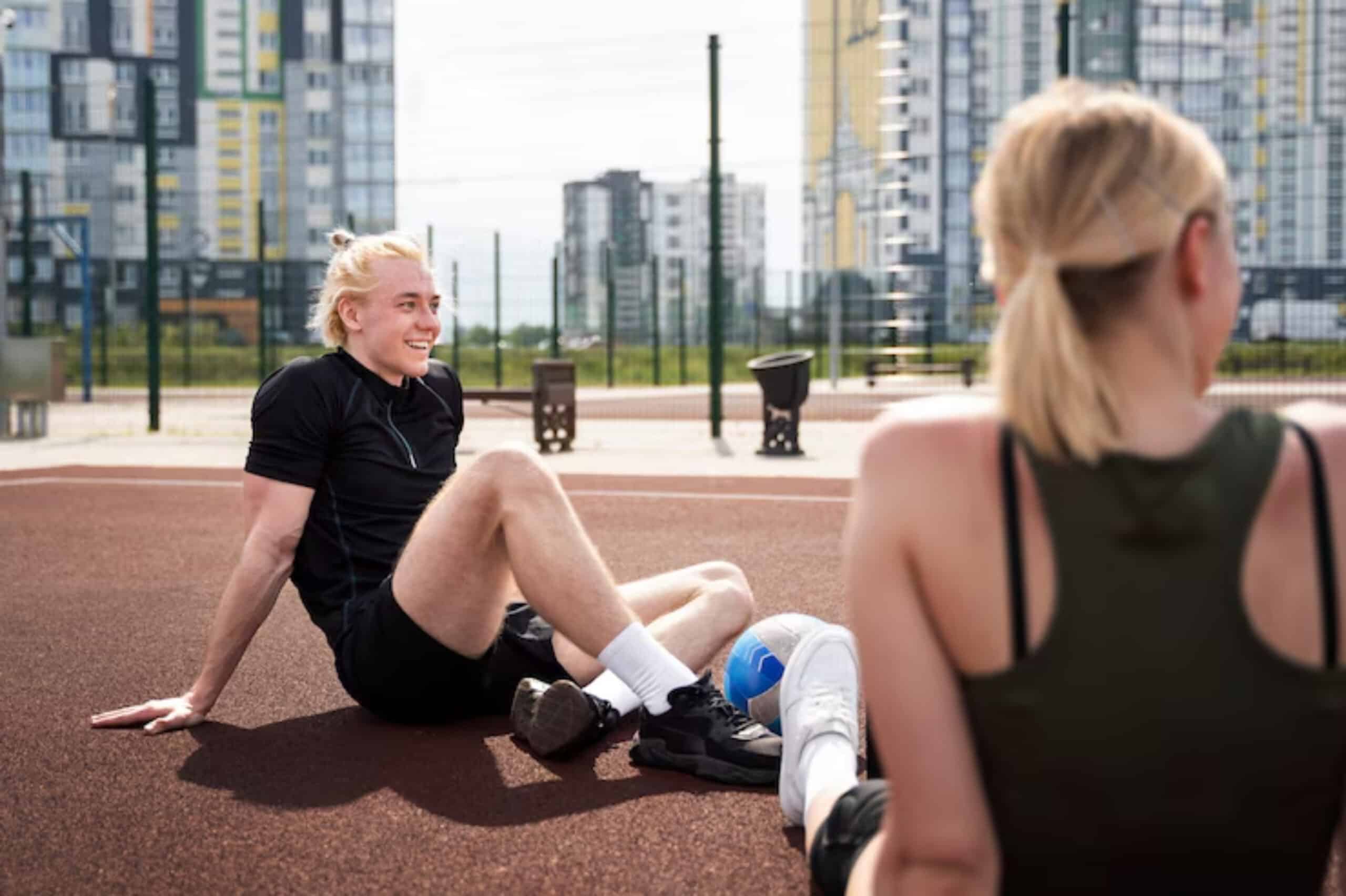 Two individuals seated on a track in an urban outdoor sports facility, with buildings and a fence in the background.