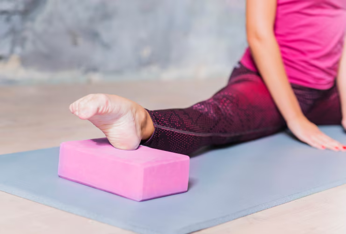 A person in athletic wear sits on a yoga mat, resting with one arm over their knee, surrounded by natural light. 