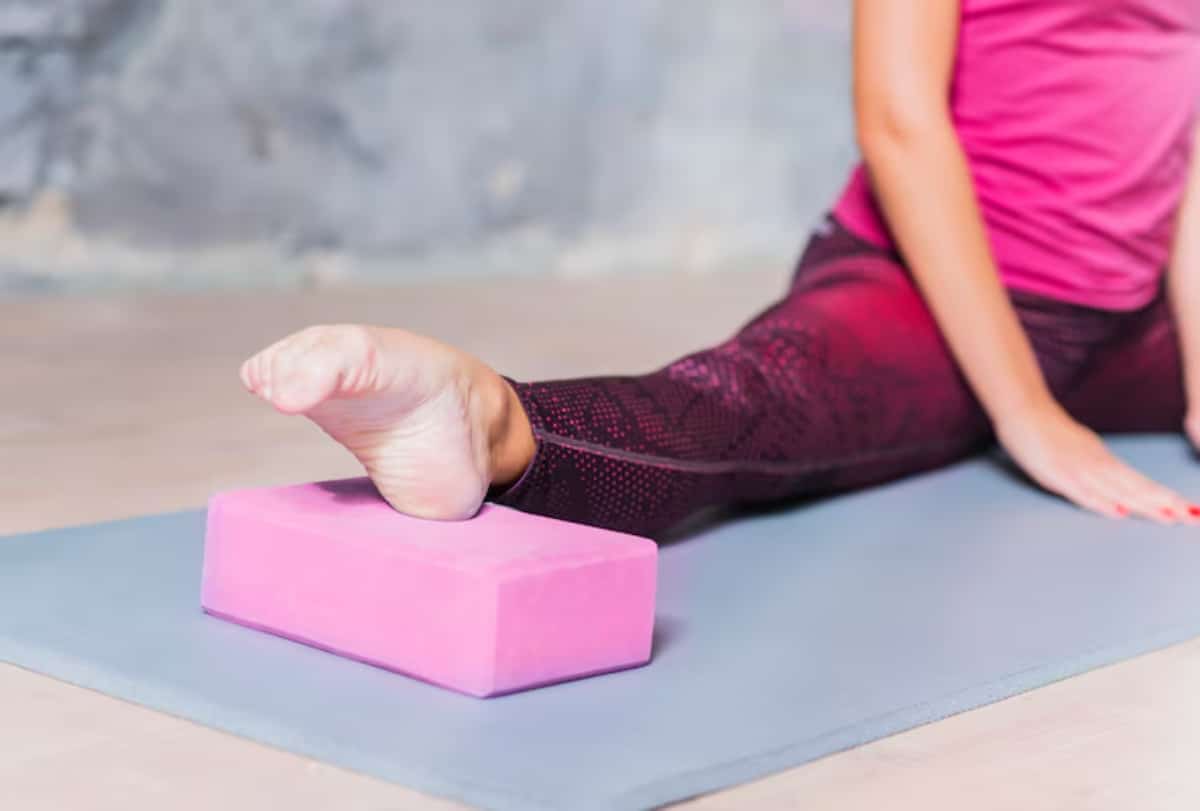 A person in athletic wear sits on a yoga mat, resting with one arm over their knee, surrounded by natural light.