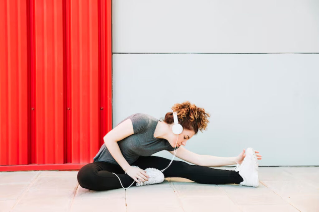 A person in a gray shirt and black leggings stretching on a tiled floor near a vibrant red wall, wearing headphones.