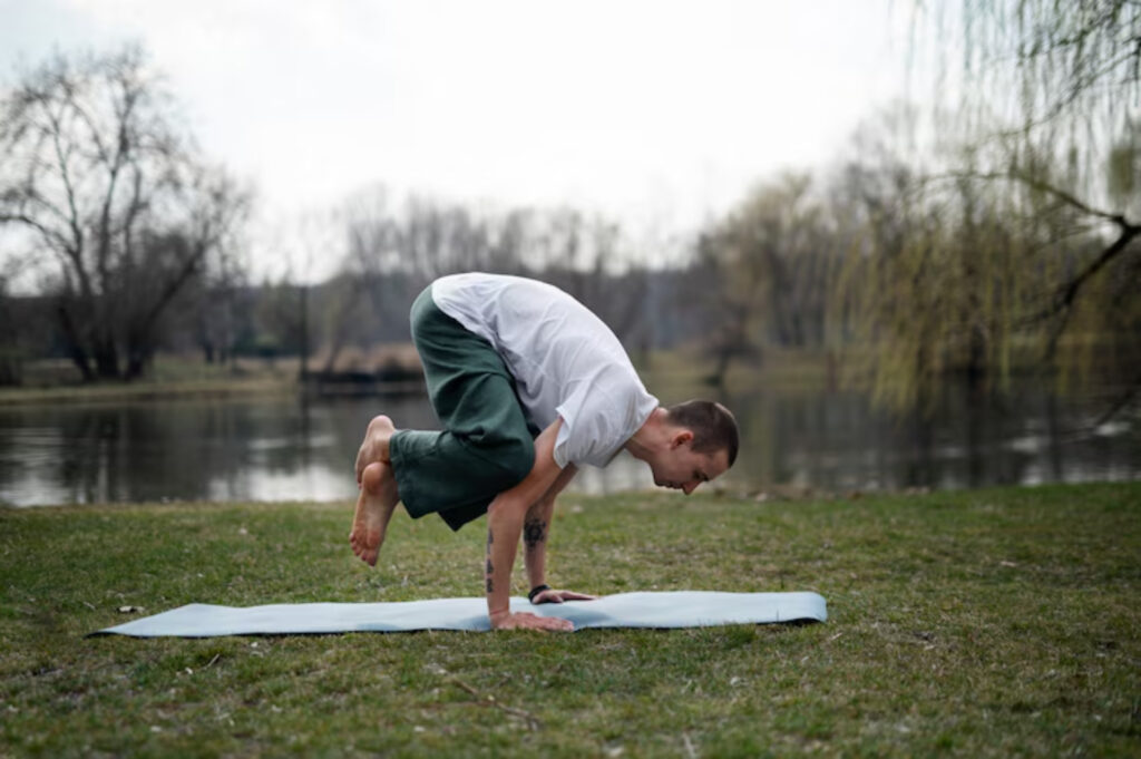 A person practicing yoga on a mat by a serene lake, performing a challenging pose, surrounded by trees and grass.