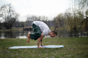 A person practicing yoga on a mat by a serene lake, performing a challenging pose, surrounded by trees and grass.
