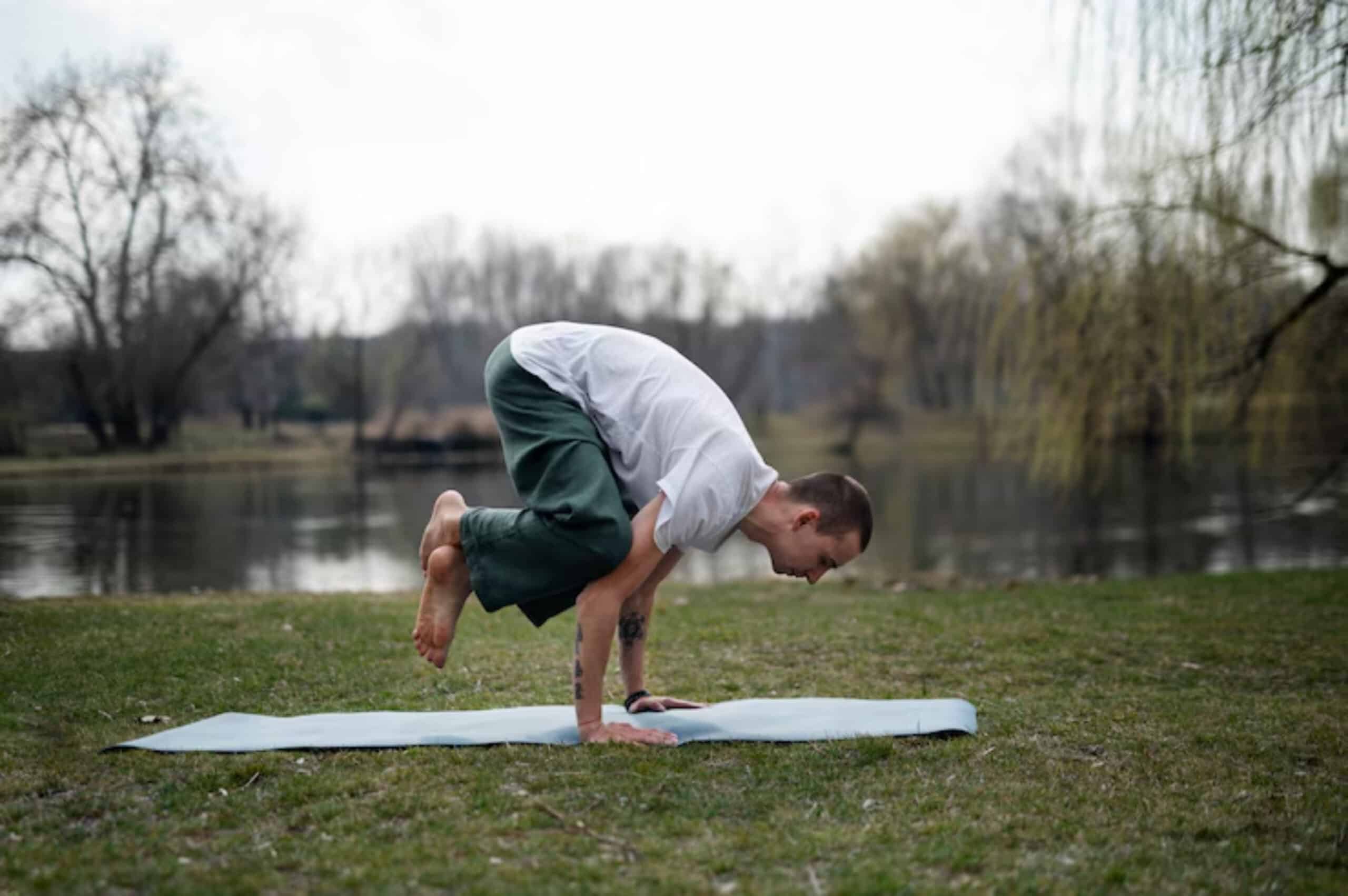A person practicing yoga on a mat by a serene lake, performing a challenging pose, surrounded by trees and grass.