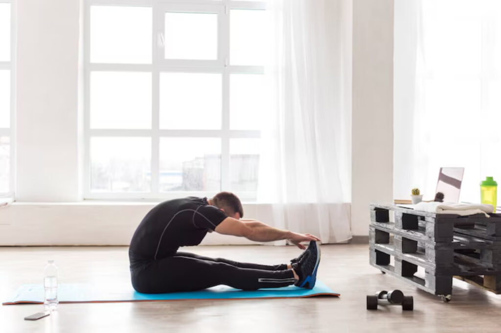 A man in athletic wear stretches on a blue yoga mat in a bright, minimalist room with windows, water bottle, and dumbbell nearby.