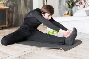 A person in black athletic wear stretches on a yoga mat in a bright, modern room with weights and a bed in the background.
