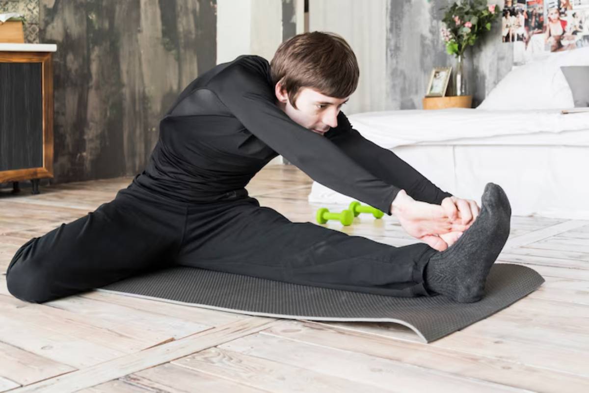 A person in black athletic wear stretches on a yoga mat in a bright, modern room with weights and a bed in the background.