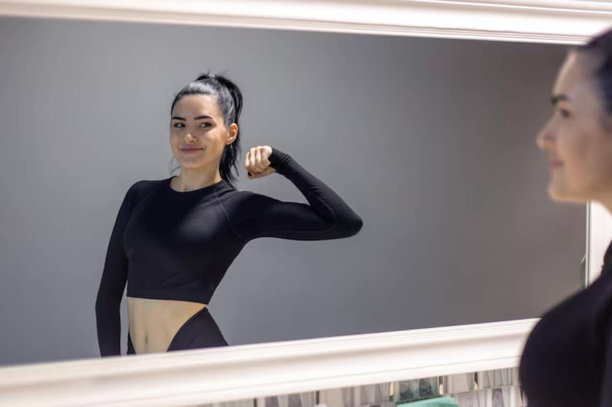 A person in a black athletic outfit flexes their arm while admiring their reflection in a stylish, framed mirror.