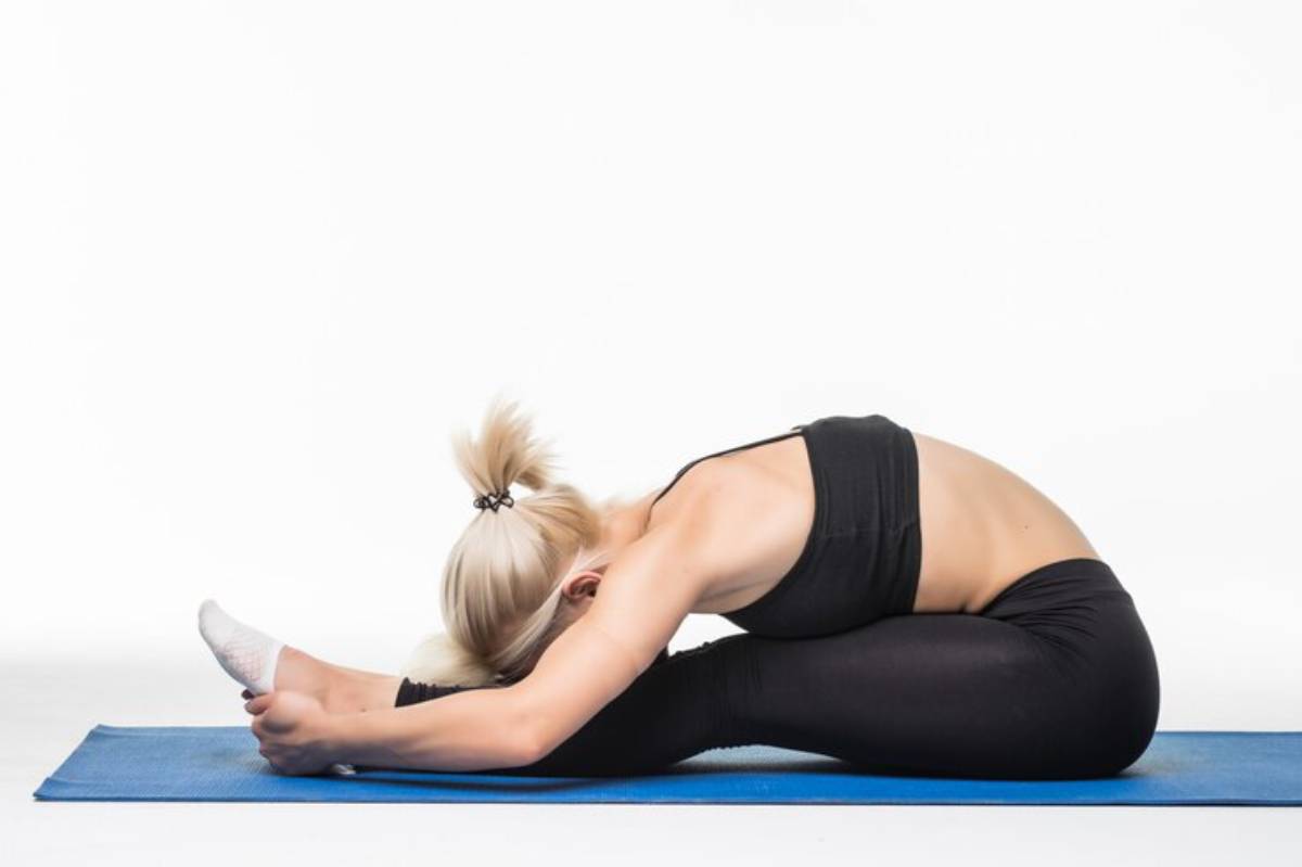 A woman with blonde hair stretches forward in a seated position on a blue yoga mat, practicing flexibility and relaxation.