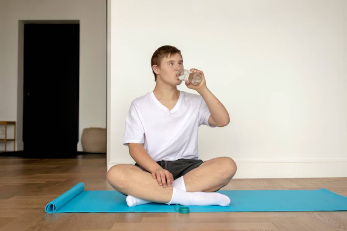A person sitting cross-legged on a blue yoga mat, sipping from a glass of water in a minimalist room. 