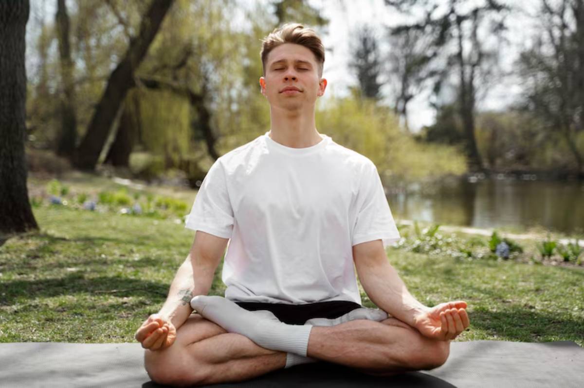 A person sits cross-legged on a yoga mat outdoors, meditating peacefully by a serene lake surrounded by greenery.