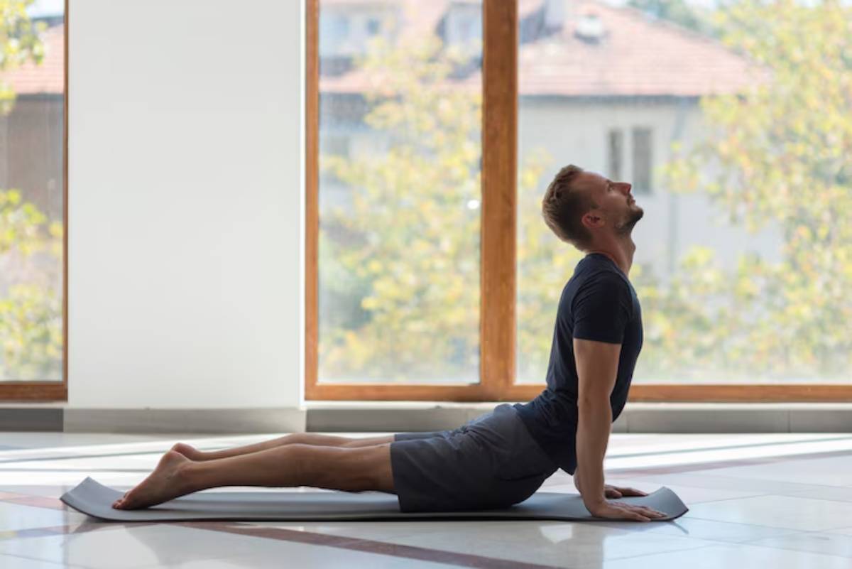  A person practices yoga in a bright room, performing the upward-facing dog pose, with large windows revealing a sunny outdoor view.