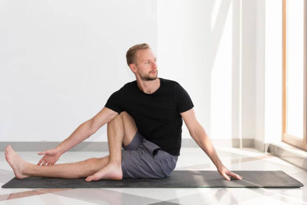 A person in a black t-shirt and gray shorts practices a seated yoga stretch on a mat, surrounded by bright, natural light.